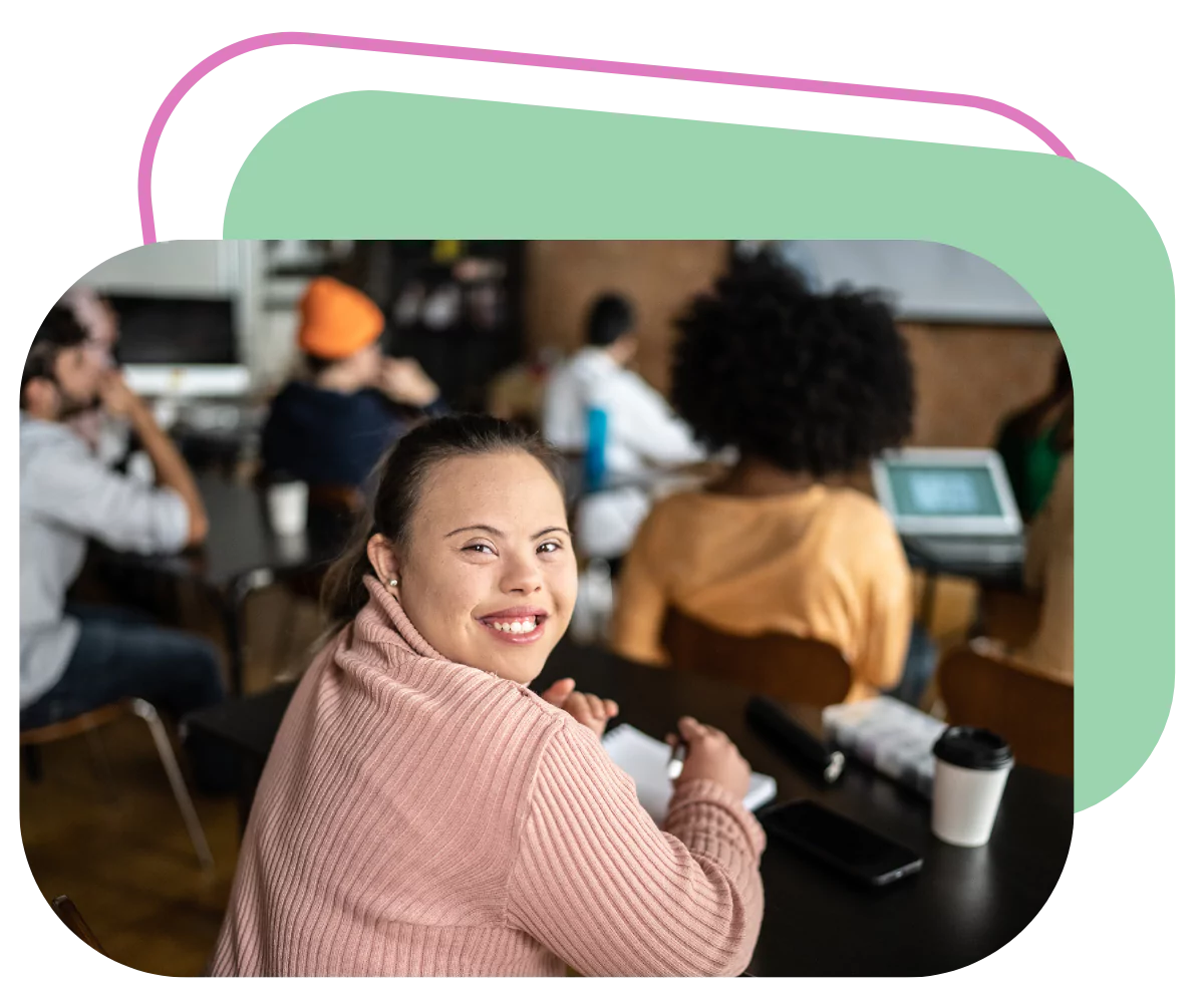 A down syndrome woman is sitting at a university lecture taking notes. She's smiling at the camera
