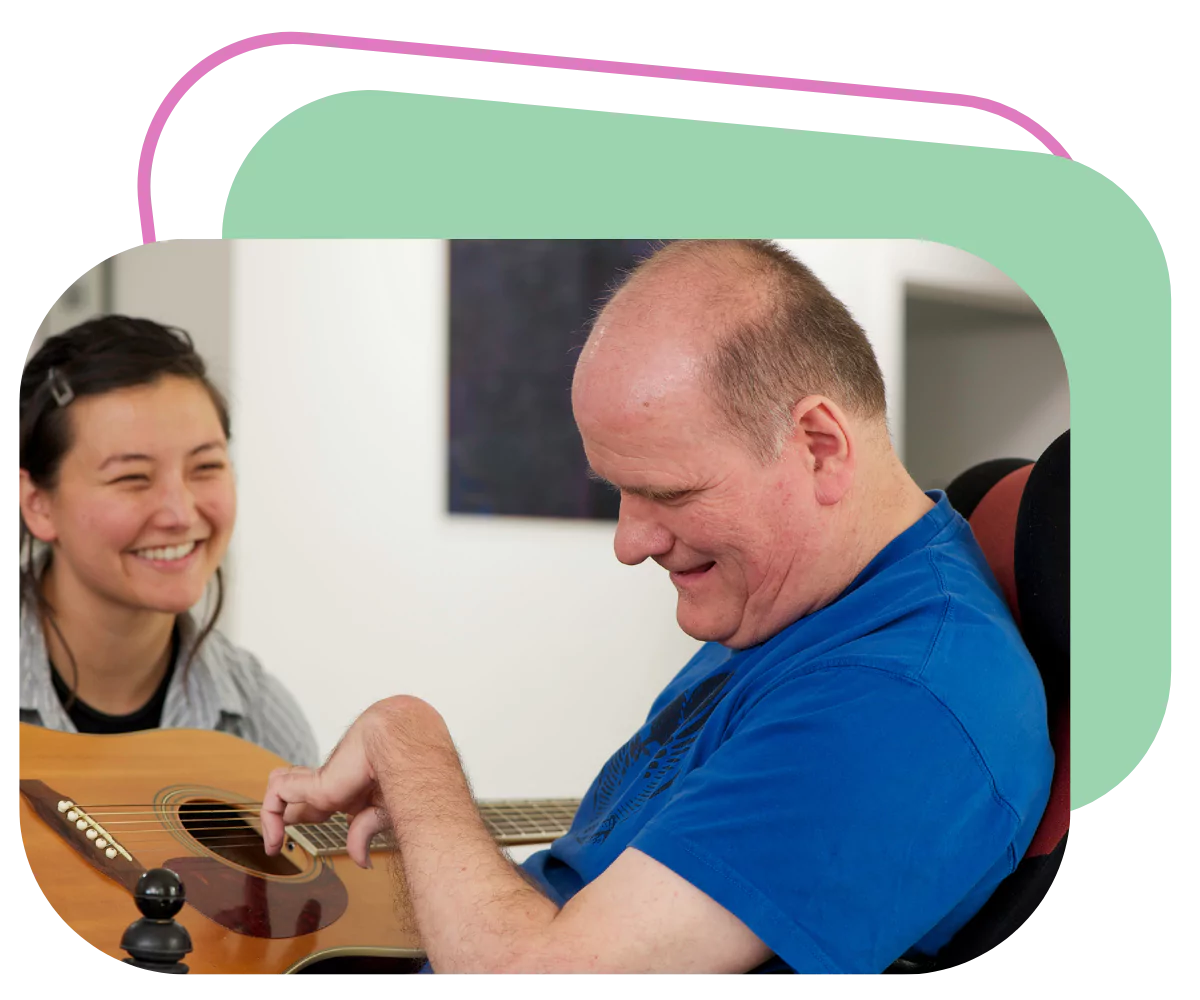 A disabled man is holding and learning to play a guitar. Hi caregiver is smiling besides him in encouragement